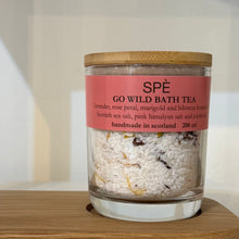A glass jar candle or bath product labeled 'SPÉ GO WILD BATH TEA' with a pink label and bamboo lid, sitting on a wooden surface against a soft beige background.