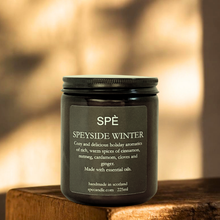 A dark glass jar candle labeled 'SPÈ SPEYSIDE WINTER' sits on a rustic wooden surface with a softly blurred warm-toned background.
