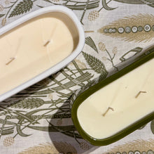 Two rectangular soy wax candles with three wicks each, one in a white container and the other in an olive green container, placed on a patterned fabric background.