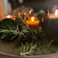 Fresh rosemary sprigs and mossy green decorative spheres arranged on a plate in a dimly lit, cozy setting with blurred candles and amber bottles in the background.