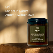 A candle jar labeled “SPÉ INISH” by Shelter in Nature, placed on a wooden surface against a warm blurred background. White text on the left reads “SPÉ Shelter in Nature Autumn | Winter 2026.