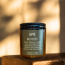 A SPÈ BOTHY candle in a dark amber glass jar with a black lid and a dark olive-green label, resting on a rustic wooden block against a warm, sunlit, blurred background.