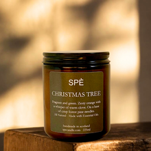 An amber glass jar candle labeled 'SPÉ Christmas Tree' sits on a rustic wooden surface with a softly blurred warm-toned outdoor background.