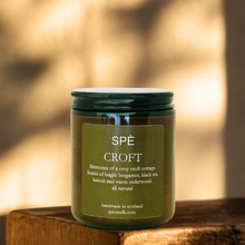 A glass jar candle with a dark green lid and olive green label reading 'SPÉ CROFT' sits on a rustic wooden surface with a softly blurred warm-toned background.