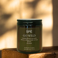 A dark green glass jar candle with a black lid and a muted olive-green label reading 'SPÉ GO WILD' sits on a rustic wooden surface with a softly blurred warm-toned background.