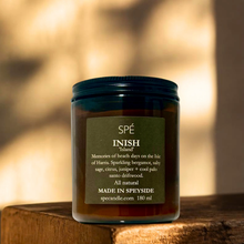A SPÉ INISH 'Island' scented candle in a dark amber glass jar with a dark green label, resting on a rustic wooden surface against a warm, sunlit, blurred background.