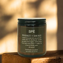 A candle in a dark glass jar with a black lid and a dark label reading 'SPÉ WHISKY + SMOKE', placed on a wooden surface with a softly blurred warm-toned background.