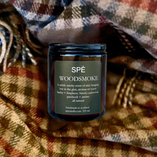A dark glass jar candle labeled 'SPÉ WOODSMOKE' sits on a plaid blanket with brown, beige, and blue tones. The candle jar is centered, and the label details the scent notes of misty hanging pine, birch, barley, and fireplace smoke.